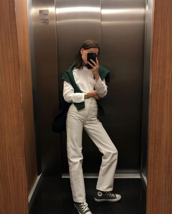 Cô gái diện toàn bộ trang phục màu trắng gồm áo thun và quần ống rộng, khoác hờ áo len màu kem trên vai, phong cách thanh lịch