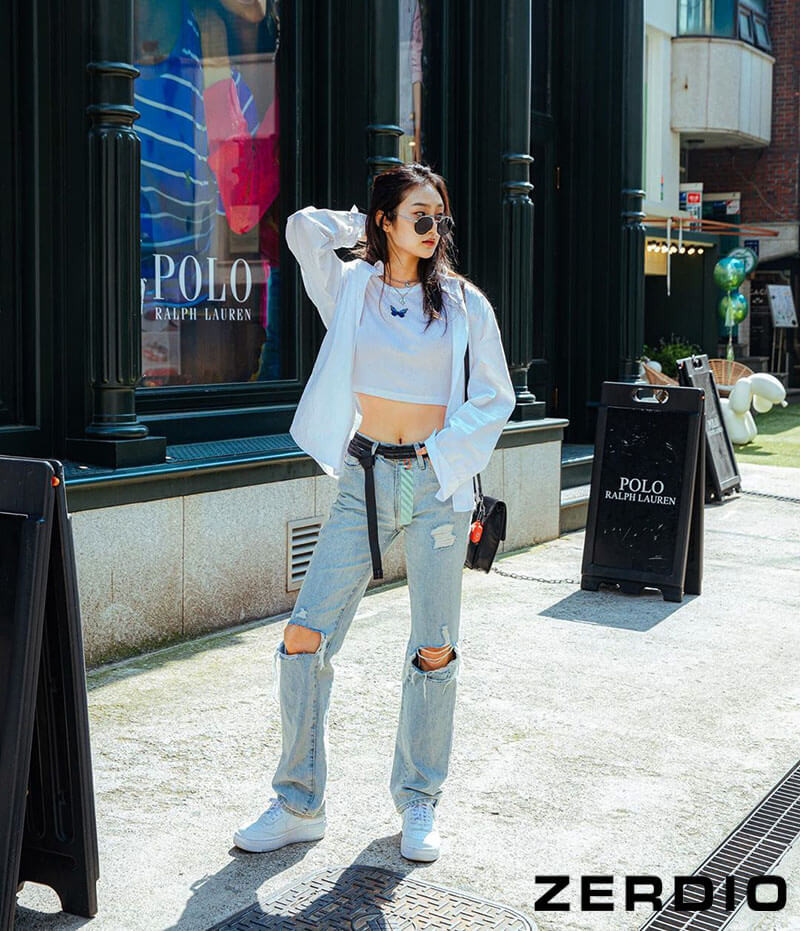 Cô gái trẻ cá tính trong quần jeans rách ống rộng, áo thun trắng và áo khoác denim, hoàn thiện phong cách hiphop nữ đường phố.